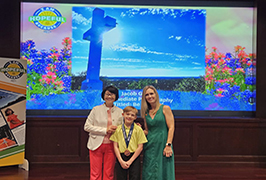  A woman, a boy, a woman with a picture of a large cross with the sun on top of it behind them.